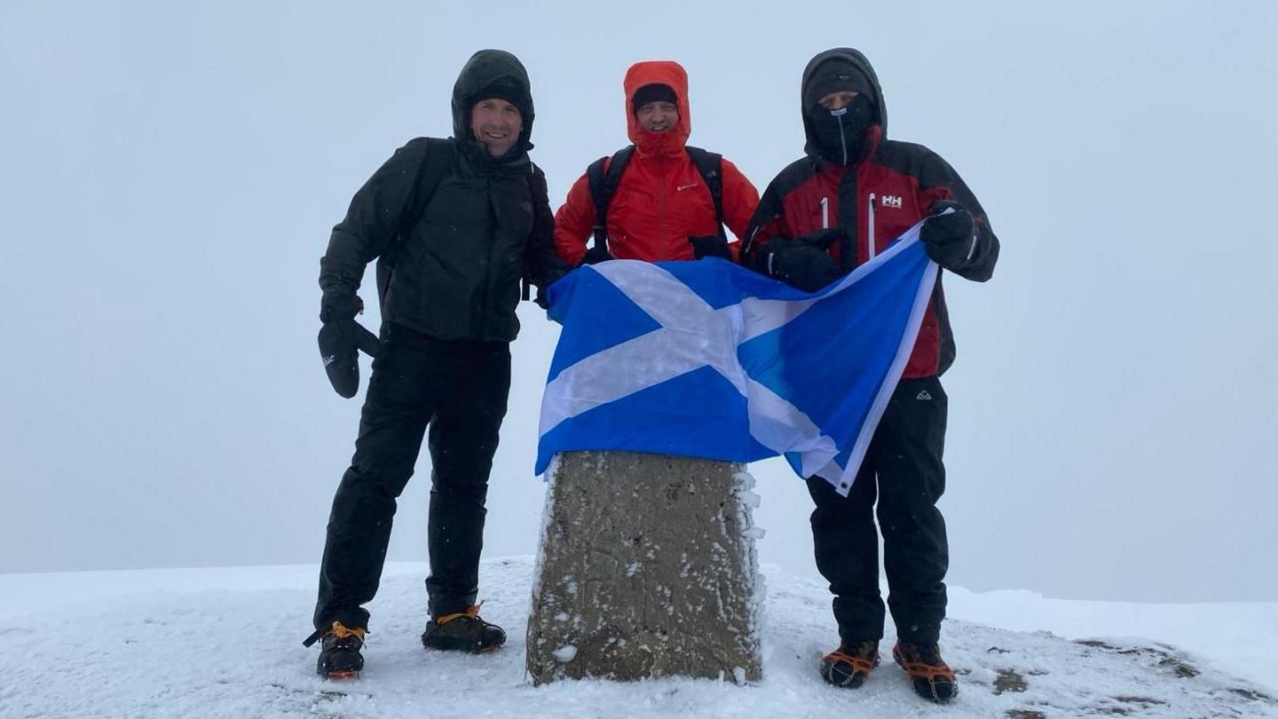 Three men wearing thick coats and gloves stood in snow holding a blue and white Scottish flag.