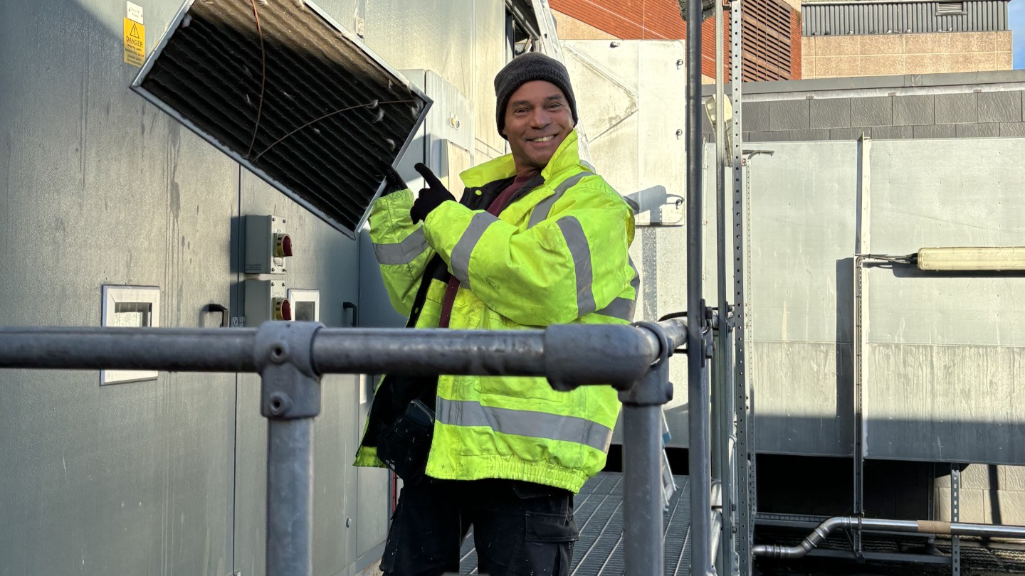 Mike Elliot wearing black cargo trousers, a yellow hi-vis coat, brown beanie hat and black gloves. He is standing on a metal scaffolding platform on the side of the Bristol Old Vic theatre, holding up what looks like a vent panel in one hand and pointing at it with the other, while smiling at the camera. 