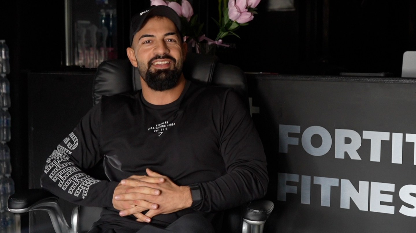 Kamal Mamedov is pictured sitting in a chair in front of a reception desk at his gym. He is wearing a black T-shirt and black trousers, while his hands sit placed in one another in his lap. He is smiling at the camera and is wearing a black cap on his head.