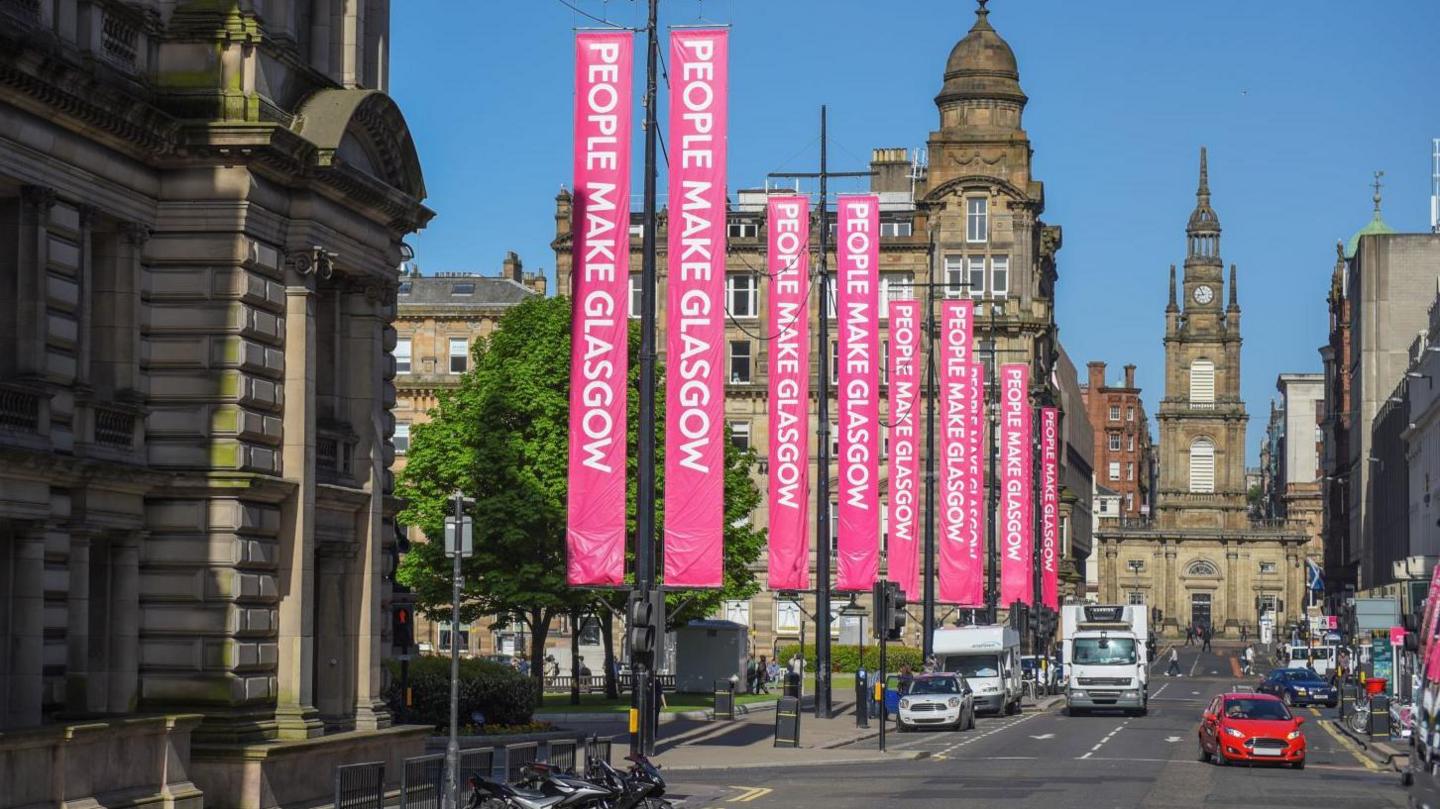 View down George Street, Glasgow with George Square on the left side and pink banners hung reading "People make Glasgow" with the St George's Tron Church on Nelson Mandela place at the very end of the street