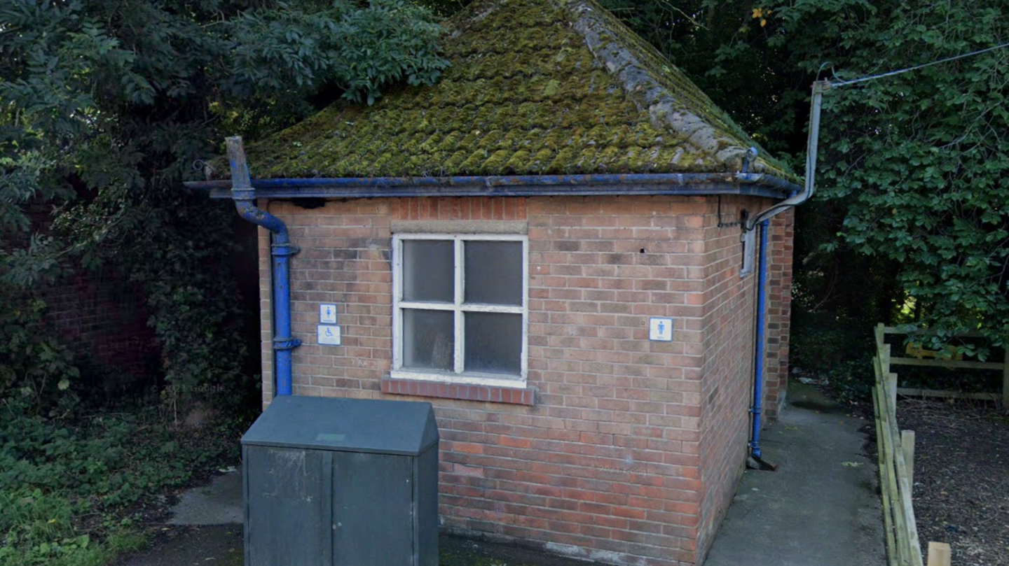 A small public toilet building with a pitched tiled roof and single square window made up of four panes. There is a sign for women and disabled people on the left and men on the right. Blue pipes run up both walls