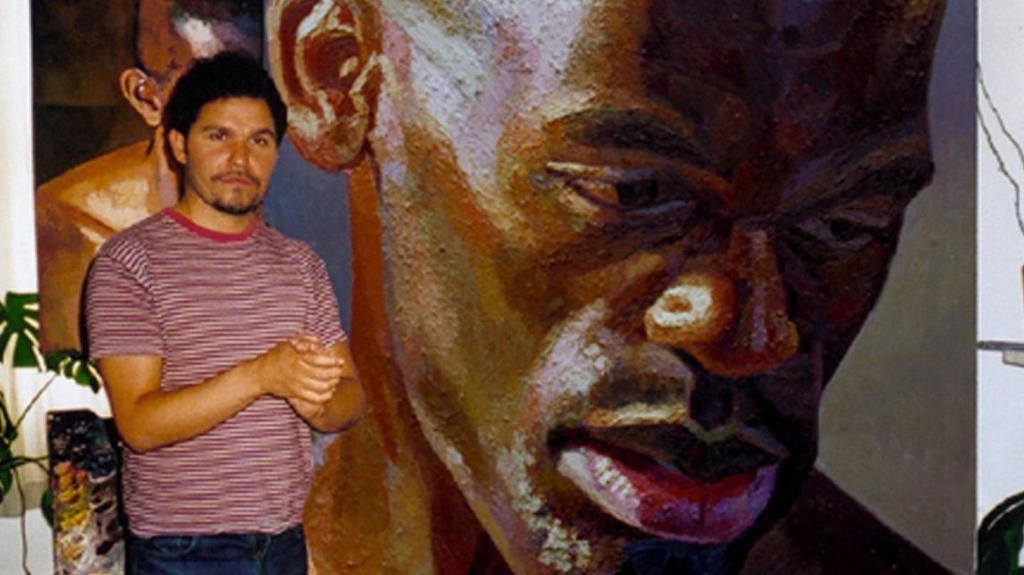 The artist stands beside a large painting of a mans face