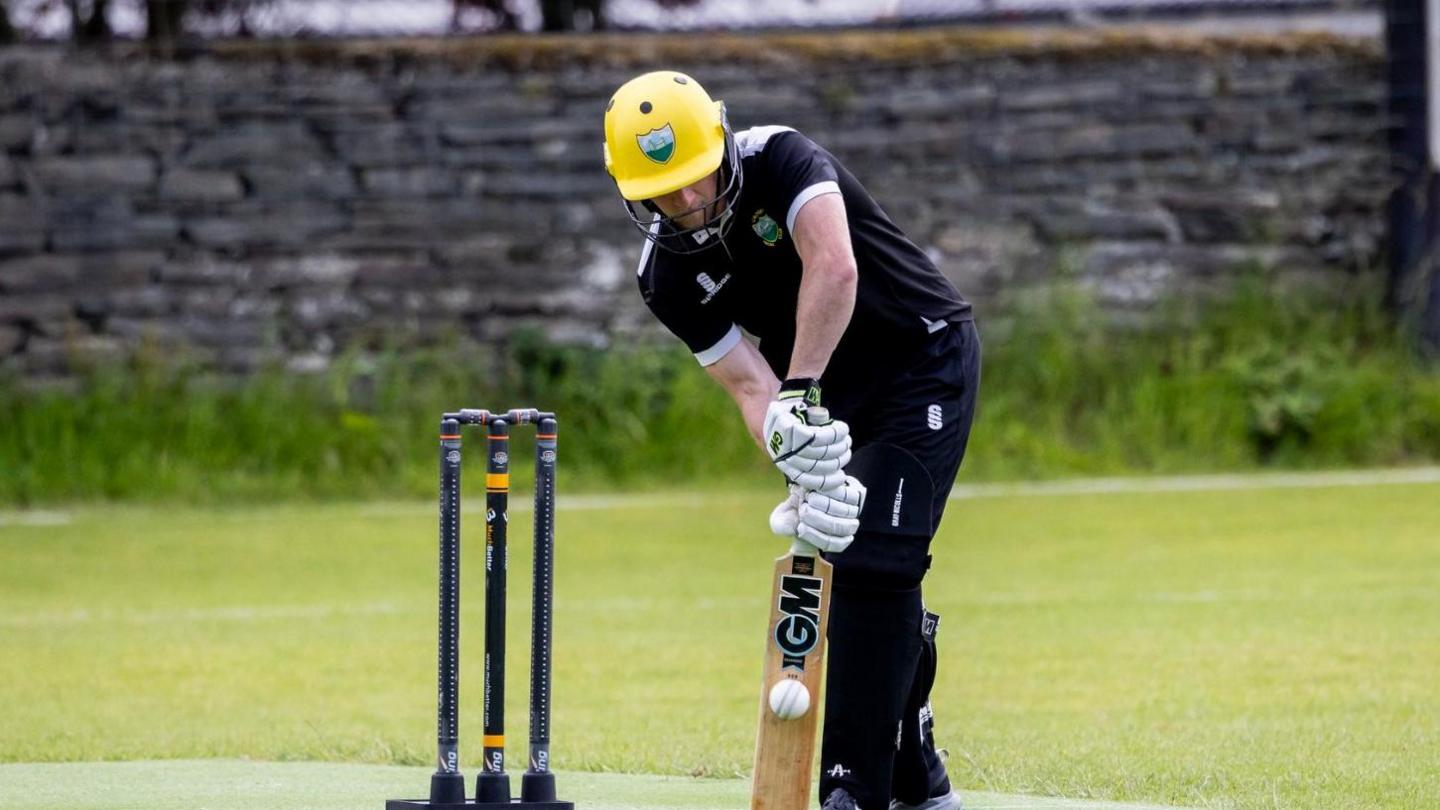 A man wears dark clothes, a yellow helmet and white gloves as he bats a ball in front of three cricket stumps.