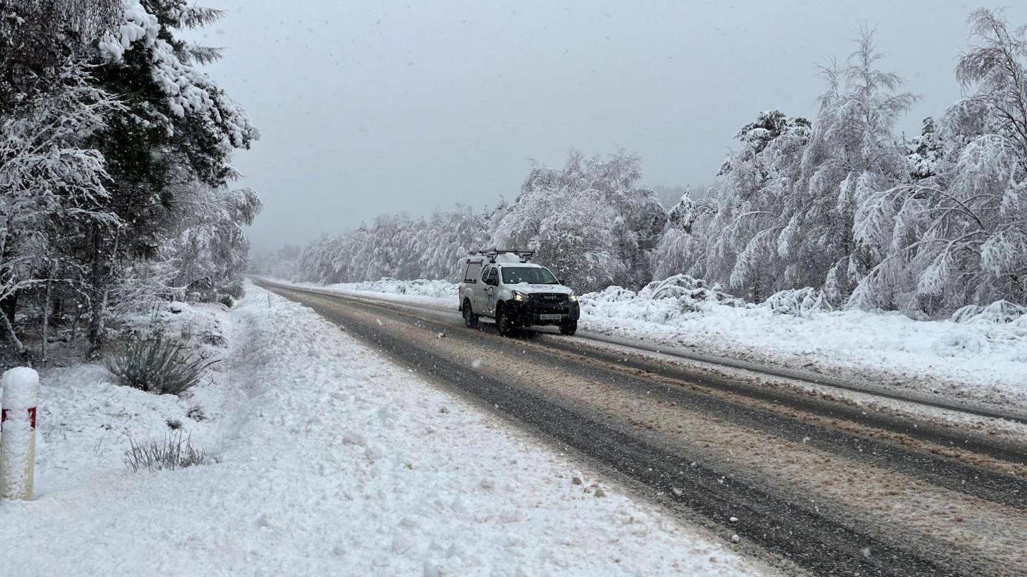 A pickup truck travels down the road which is covered in snow and slush. Trees either side of the road are covered in snow.