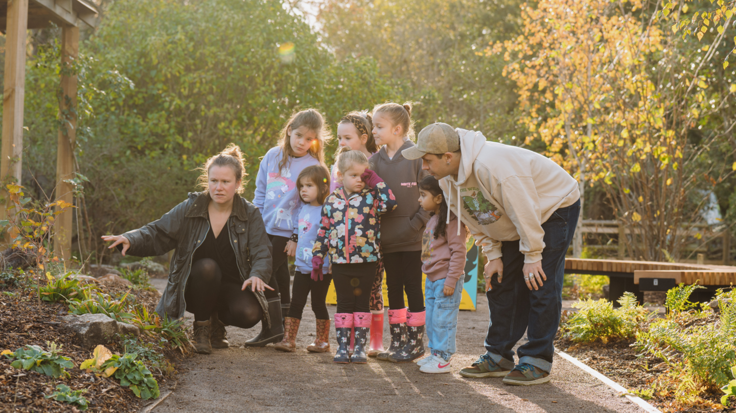Zoe, wearing a green jacket, crouched down, talking to a group of small children and showing them the garden with a male helper who is wearing a white hoodie and blue jeans.