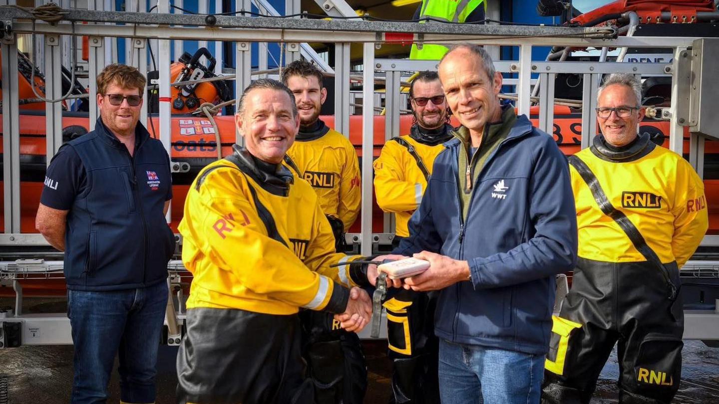Mr Tough hands over the device to Mr Murray, who has slicked back short dark hair and is wearing bright yellow RNLI waterproofs. Three volunteers in the same outfits are standing behind, with a fourth volunteer wearing a navy RNLI top and jeans. An inshore lifeboat is on a metal trailer in the background.