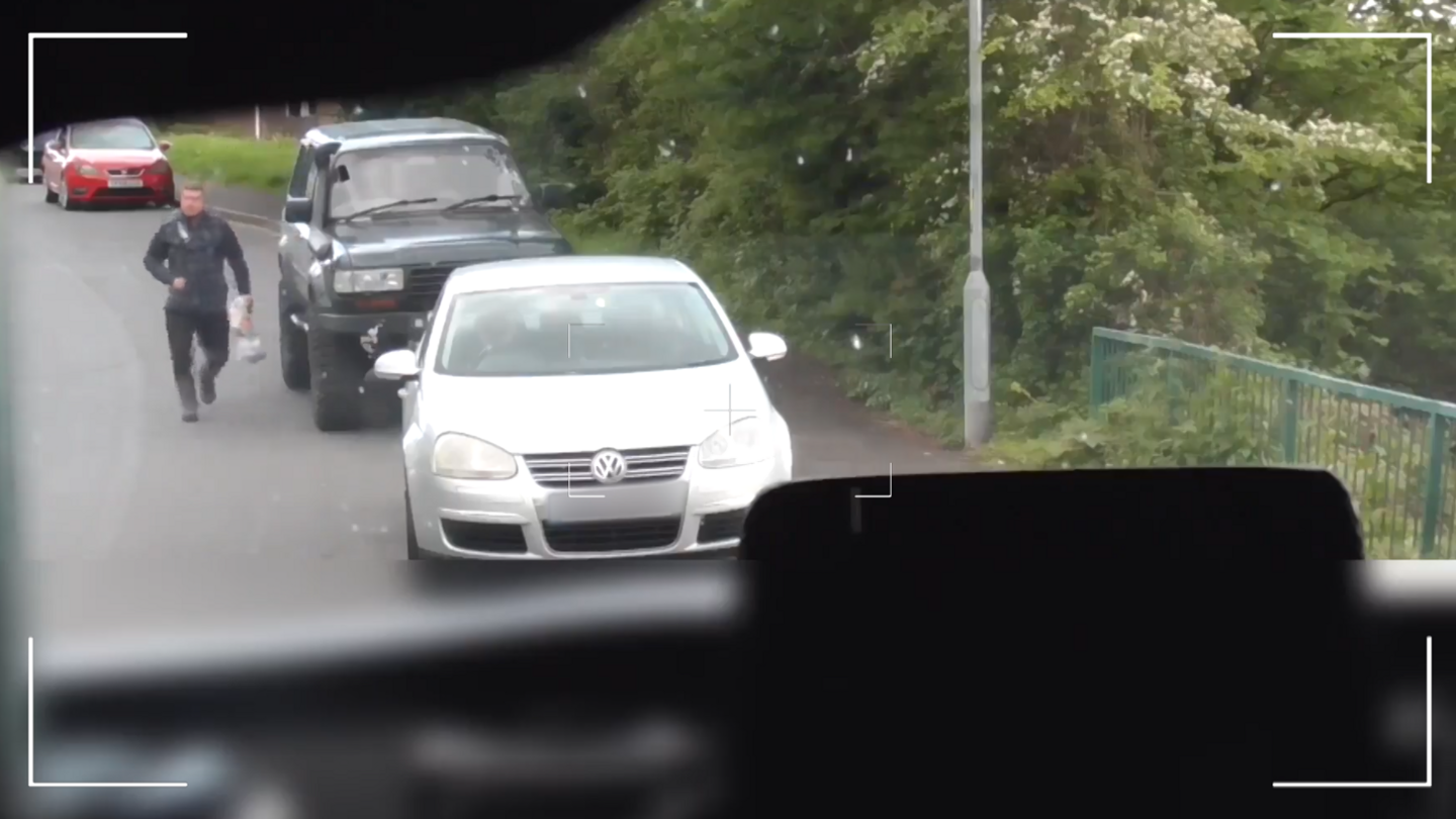 Still image of surveillance footage of a brown-haired man wearing a black coat, black shoes and black trousers walking on a road carrying a supermarket carrier bag past a black vehicle towards a silver car 