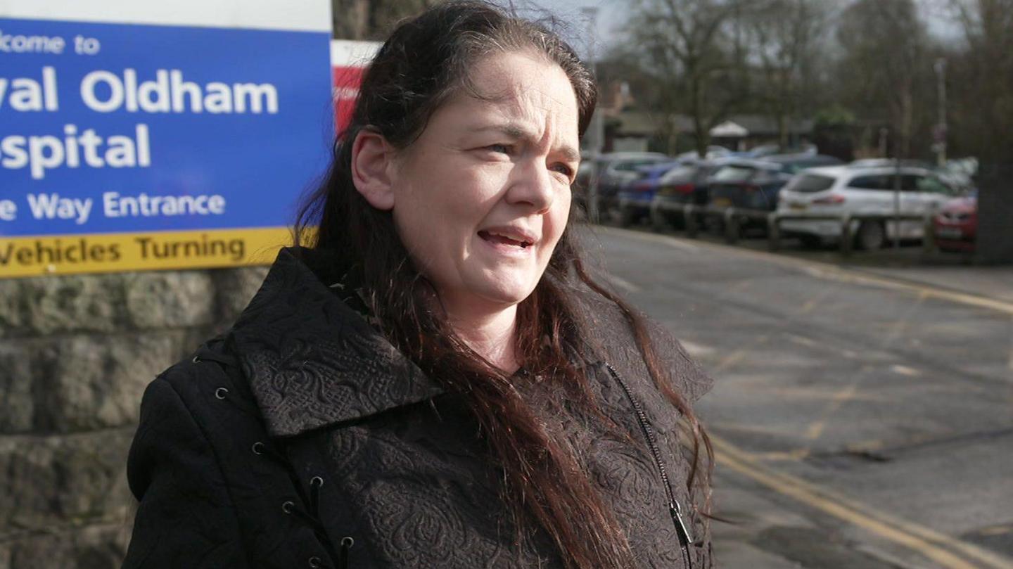 Emma Hopwood has long brown hair and a dark brown coat and is standing in front of the entrance sign at Royal Oldham Hospital.