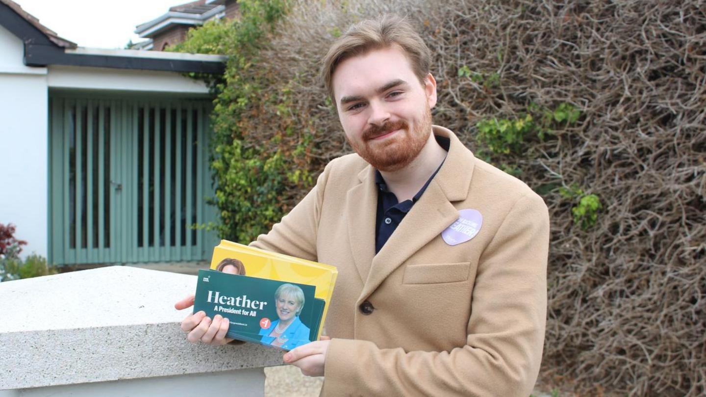 James Turley, 23, is the Chair of Dublin-Rathdown Young Fine Gael is standing outside a house. He has blonde hair and a beard. There is a hedge behind him. He is dressed in a tan overcoat over a navy polo shirt. He is holding a green flyer saying 'Heather, a president for all.' To the right of this is a picture of Heather in a blue suit jacket. He is also holding yellow flyers are seen behind.
