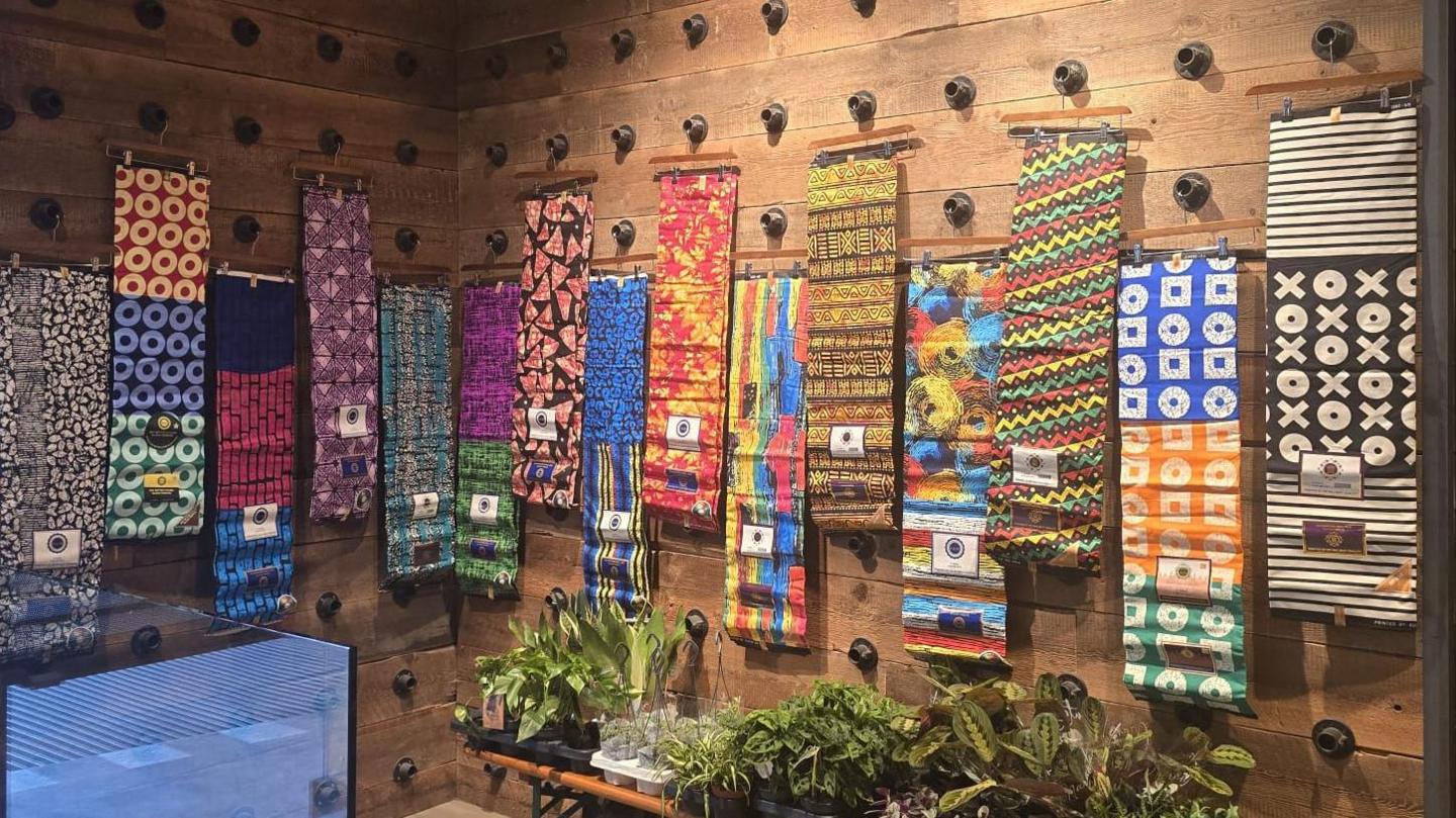 Colourful pieces of art made out of fabric were hanging on a stained brown wooden wall. There were plants standing on a wooden bench on the right.