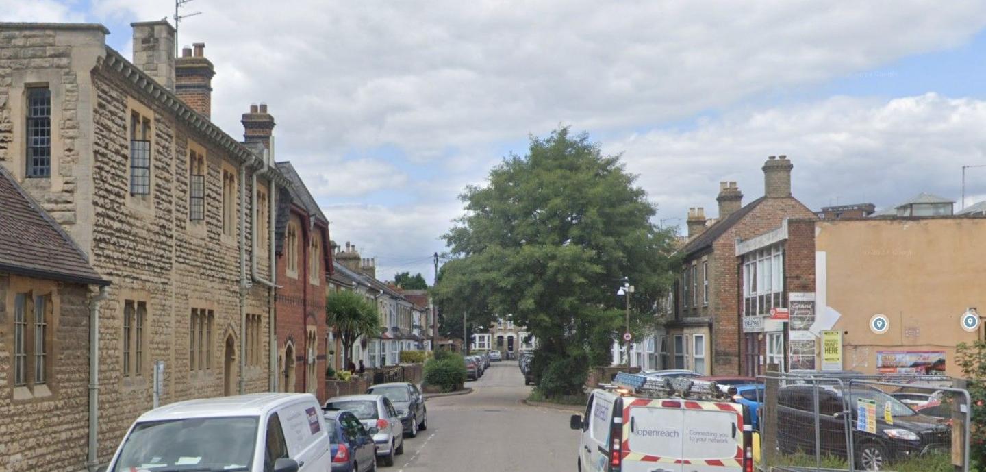 Brereton Road in Bedford with cars parked down both sides, a number of houses, a church to the left, and a tree in the distance. There are also railings to the right 