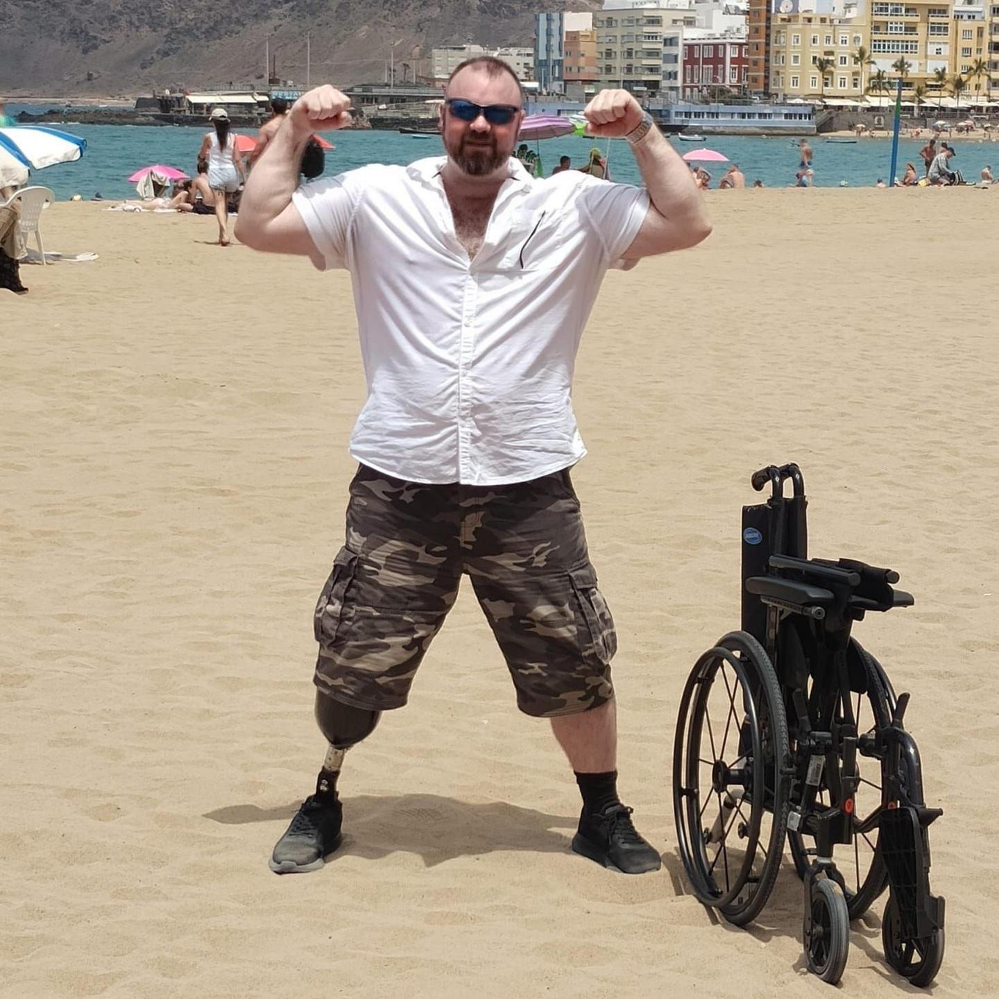 Mark Willerton standing an a beach abroad. He's wearing a prosthetic leg, is pulling a muscle man pose, showing off his large biceps. He's wearing sunglasses, a short-sleeved white shirt, and combat shorts. His wheelchair is folded up on the side.