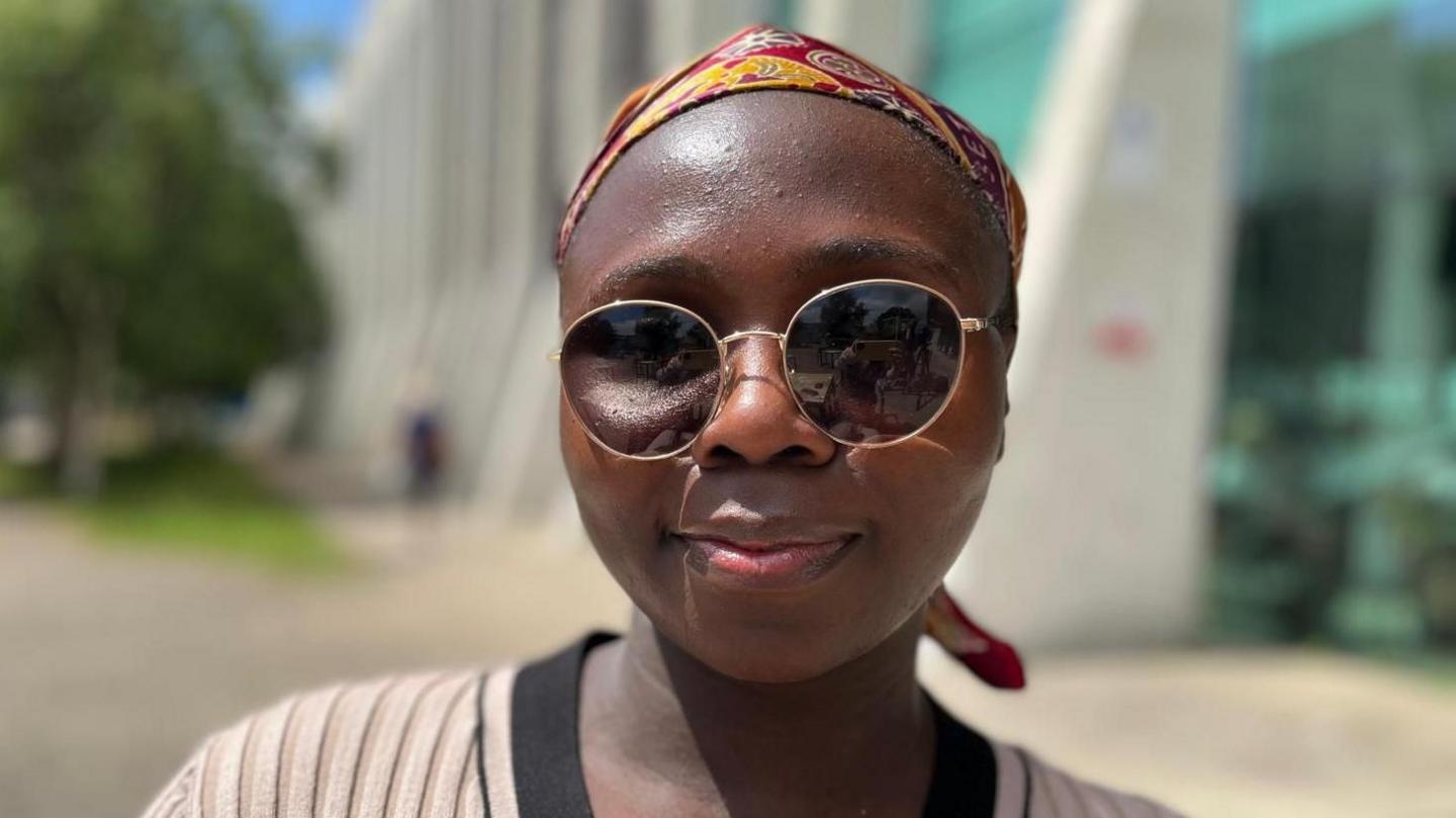 Umyima is wearing reactive glasses that have darked in the direct sunlight. She is sat outside, wearing a beige and black knitted dress and a red and yellow patterned head scarf. She has brown skin and is smiling at the camera. Behind her, slightly blurred, is a building and trees. It is a head and shoulders shot of her.