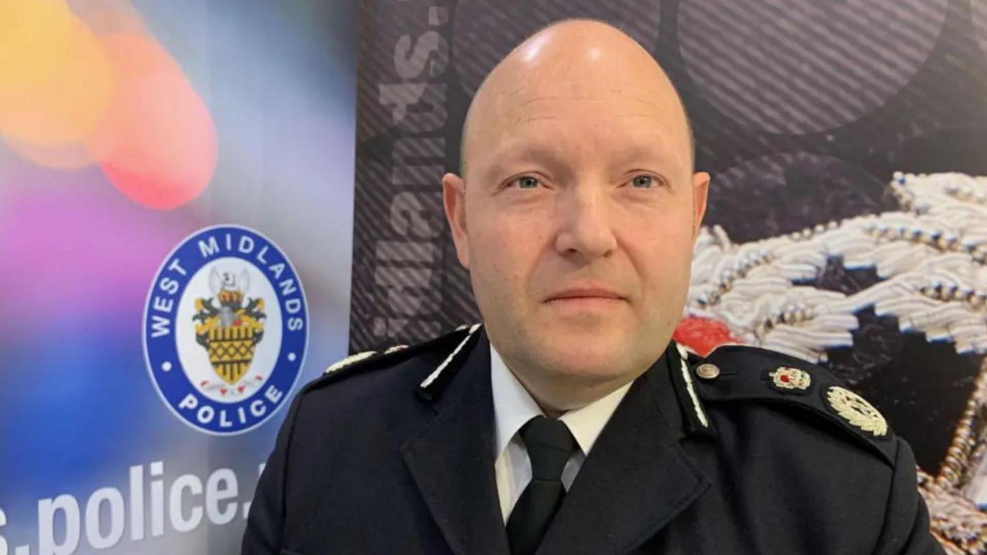 A man with a bald head in black police uniform with a blue notice behind with a police emblem on it