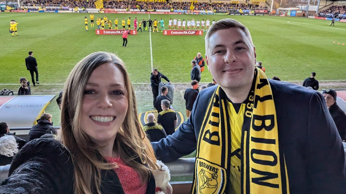 Sarah Edwards, wearing a red top and a black coat, is standing next to Jacob Collier, wearing a yellow football shirt and scarf and a black coat. They are in the spectator area of the football stadium, with two teams of players lining up on the pitch behind them.