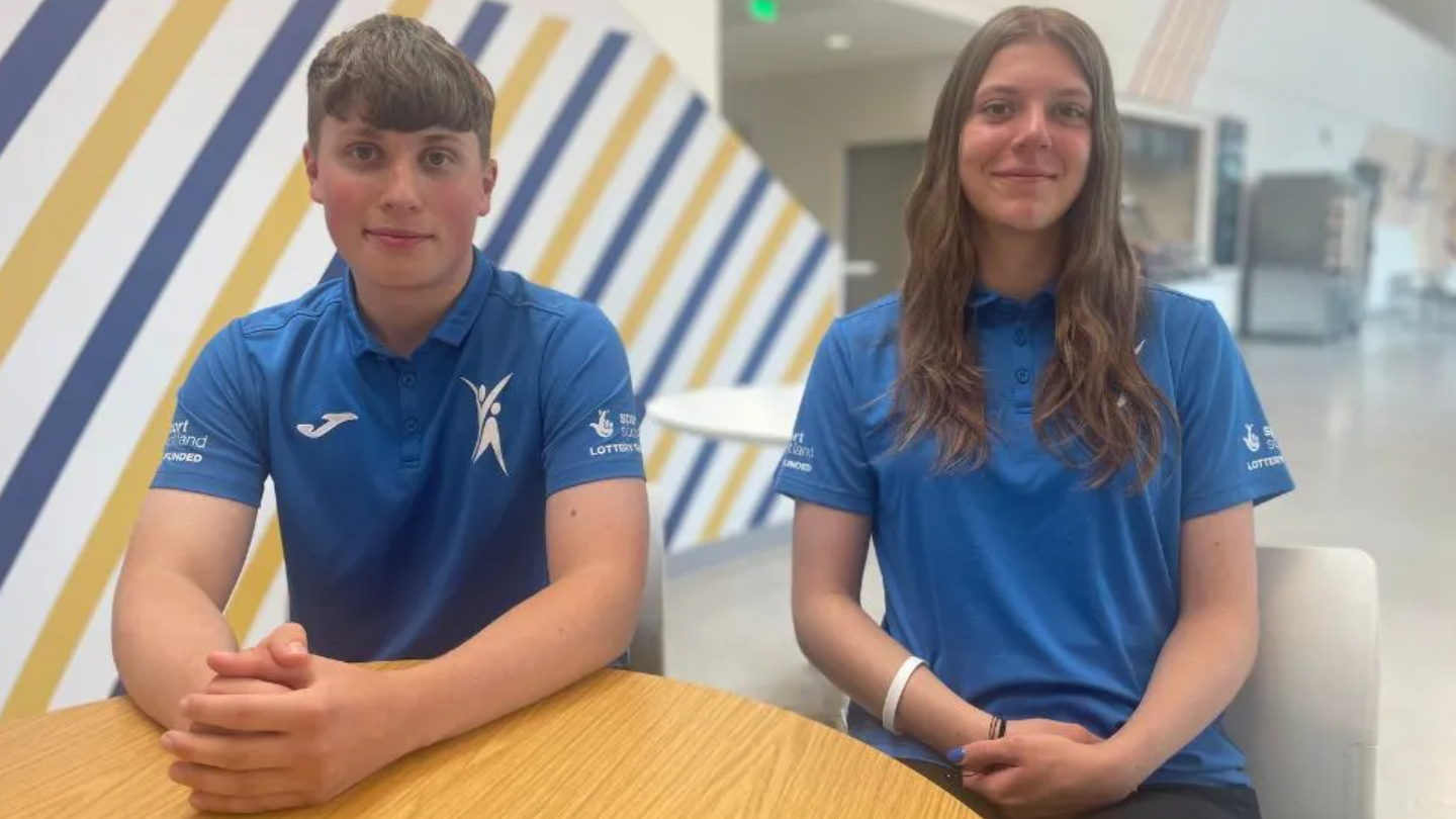 Two teenagers one with short brown hair and one with long brown hair wearing blue t-shirts smiling