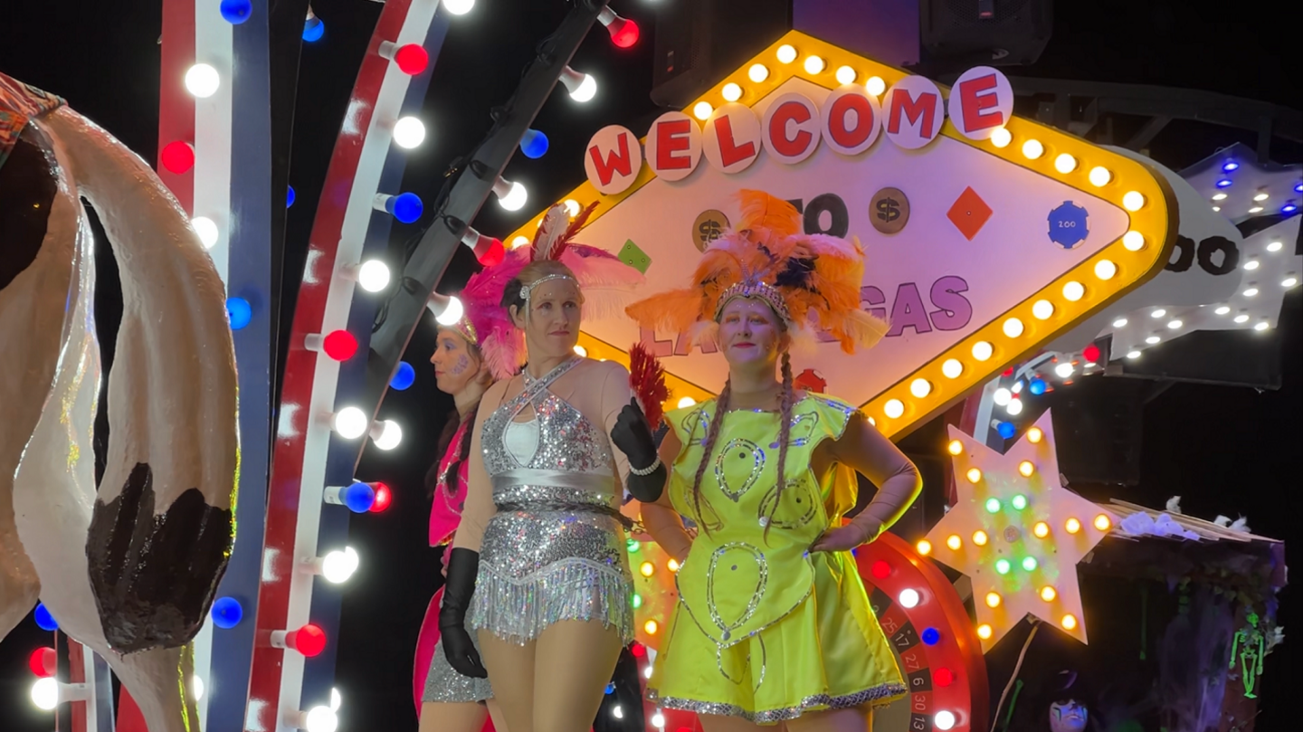 Three carnival goers on a cart, dressed in bright and sparkly clothing with feathery headpieces in front of a "Welcome to Las Vegas" sign