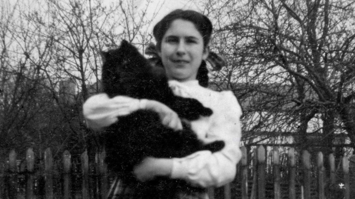 A black and white photograph of a dark-haired girl wearing a white blouse and holding a large black cat.