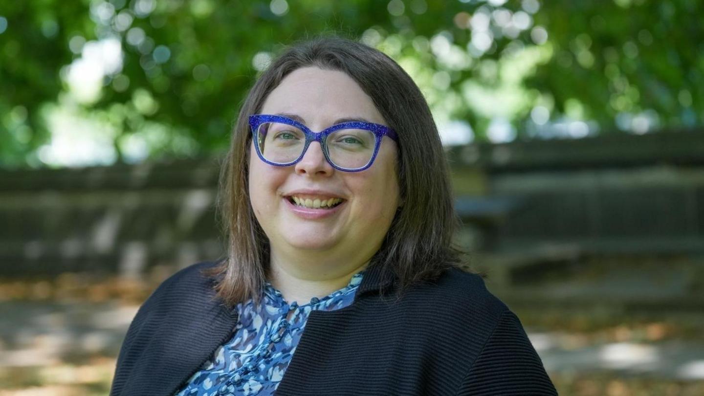 A young woman with a shoulder length bob and purple framed glasses wearing a navy jacket and blue blouse