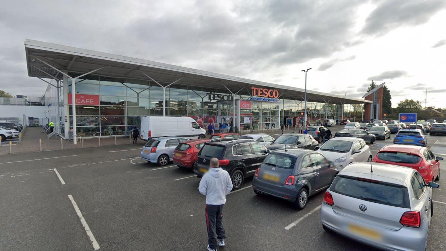 An large out of town supermarket in a modern, flat roofed building seen from a full car park with people walking in and out