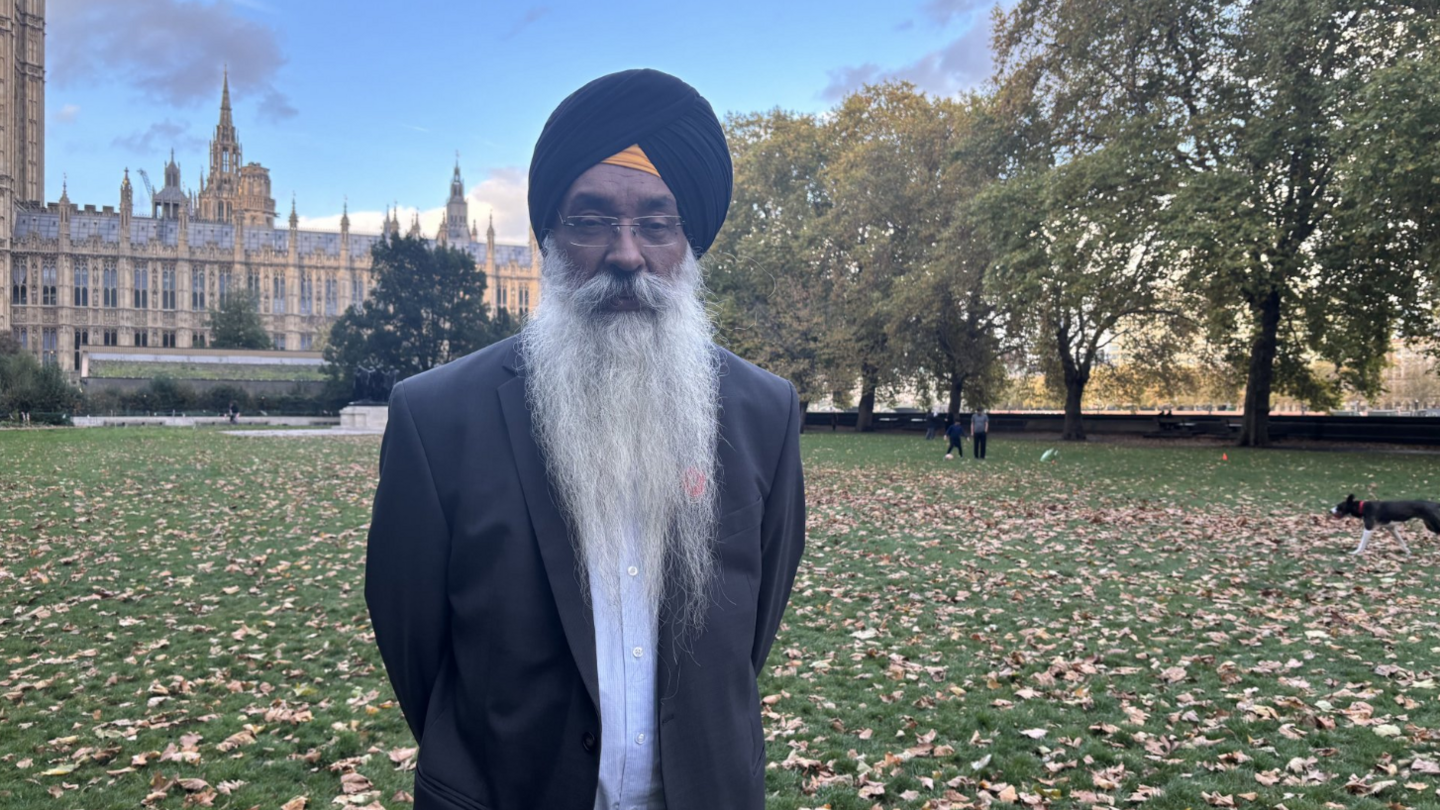 A man with a long grey beard which falls to his chest, wearing a black turban and dark suit, stands on the grass in front of the Houses of Parliament.