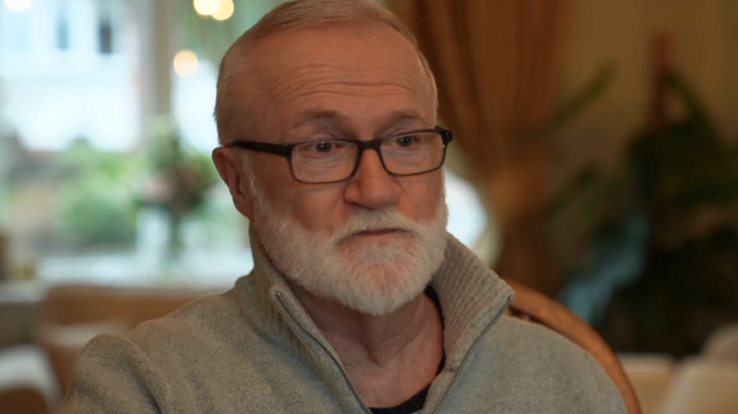 A smart looking man in his sixties, Bob Mayse. The background is blurred but appears to be his living room. He has white hair, a neat white beard, and black rectangular glasses. He wears a thick, light-grey fleece.