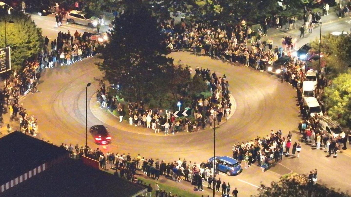 An overhead shot at night time of cars going around a roundabout whilst crowds line the pavement.