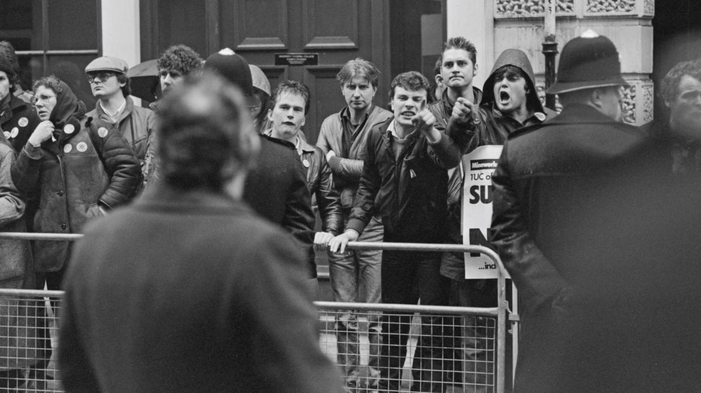 A black and white image take during the miners' strike. There are a row of angry looking men stood behind a metal fence. Two of the men are pointing their fingers at a man stood in front of them. There is a policeman stood at the side with his arms behind his back. 