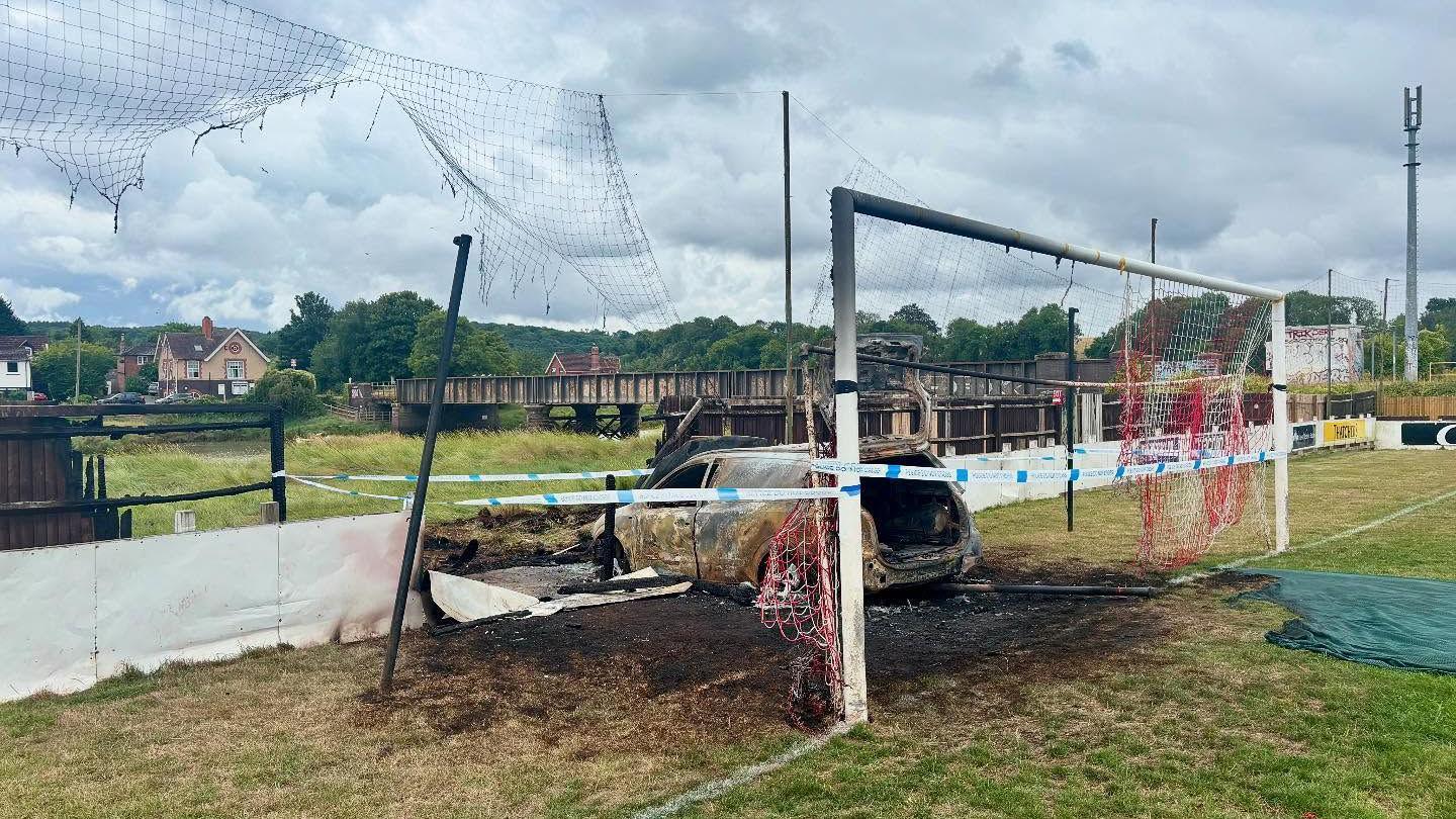 An image of a white car that has been completely burnt out, it is sitting inside a goal post on a football pitch. The goal post and its net are badly damaged, and a patch of grass around the vehicle scorched. The hoarding around the pitch is also damaged where the car has driven into it.