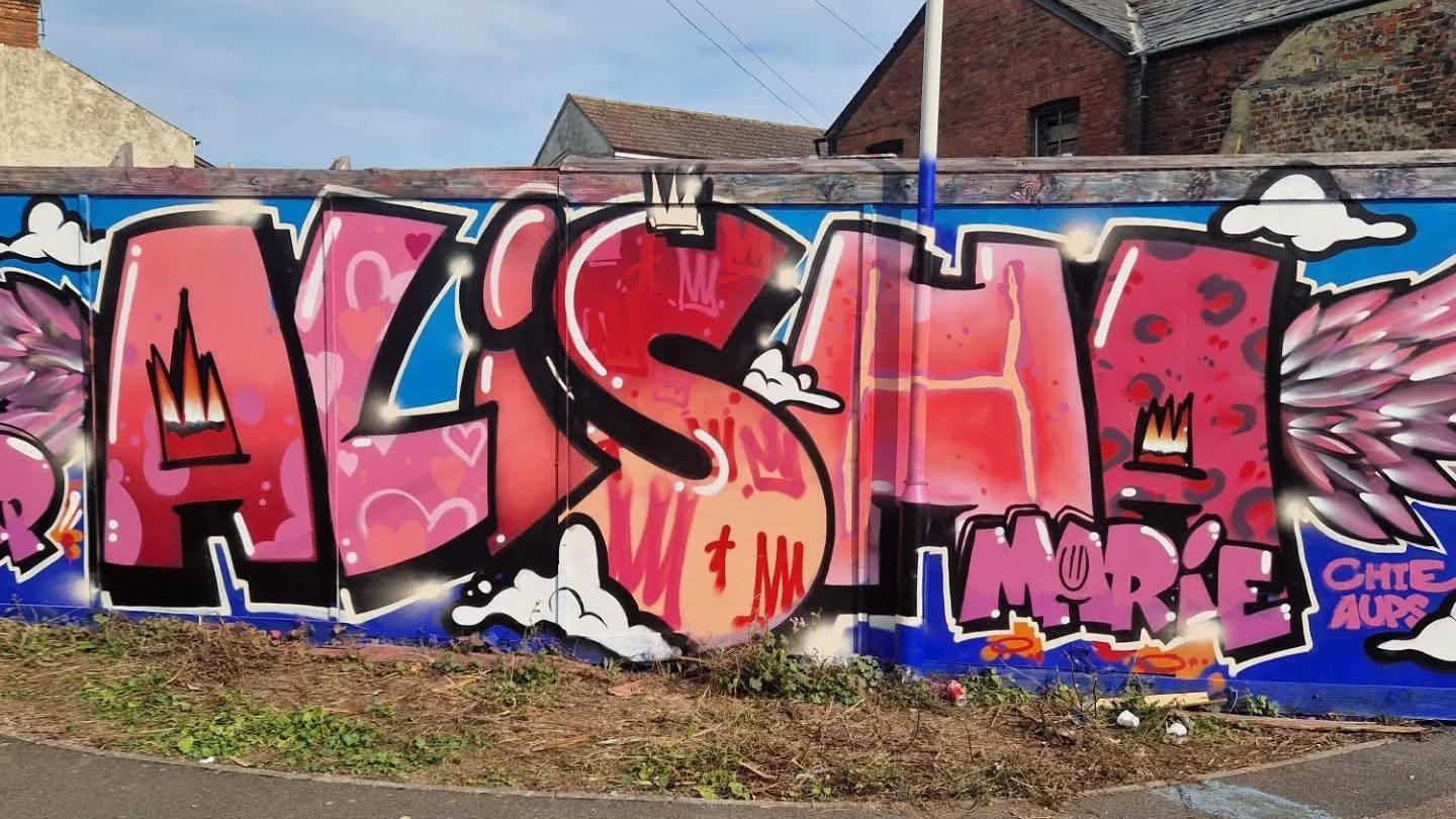 A large colourful graffiti-style mural displays the name Alisha Marie in pink and red lettering with decorative artwork and wing motifs on a wall beside a road.