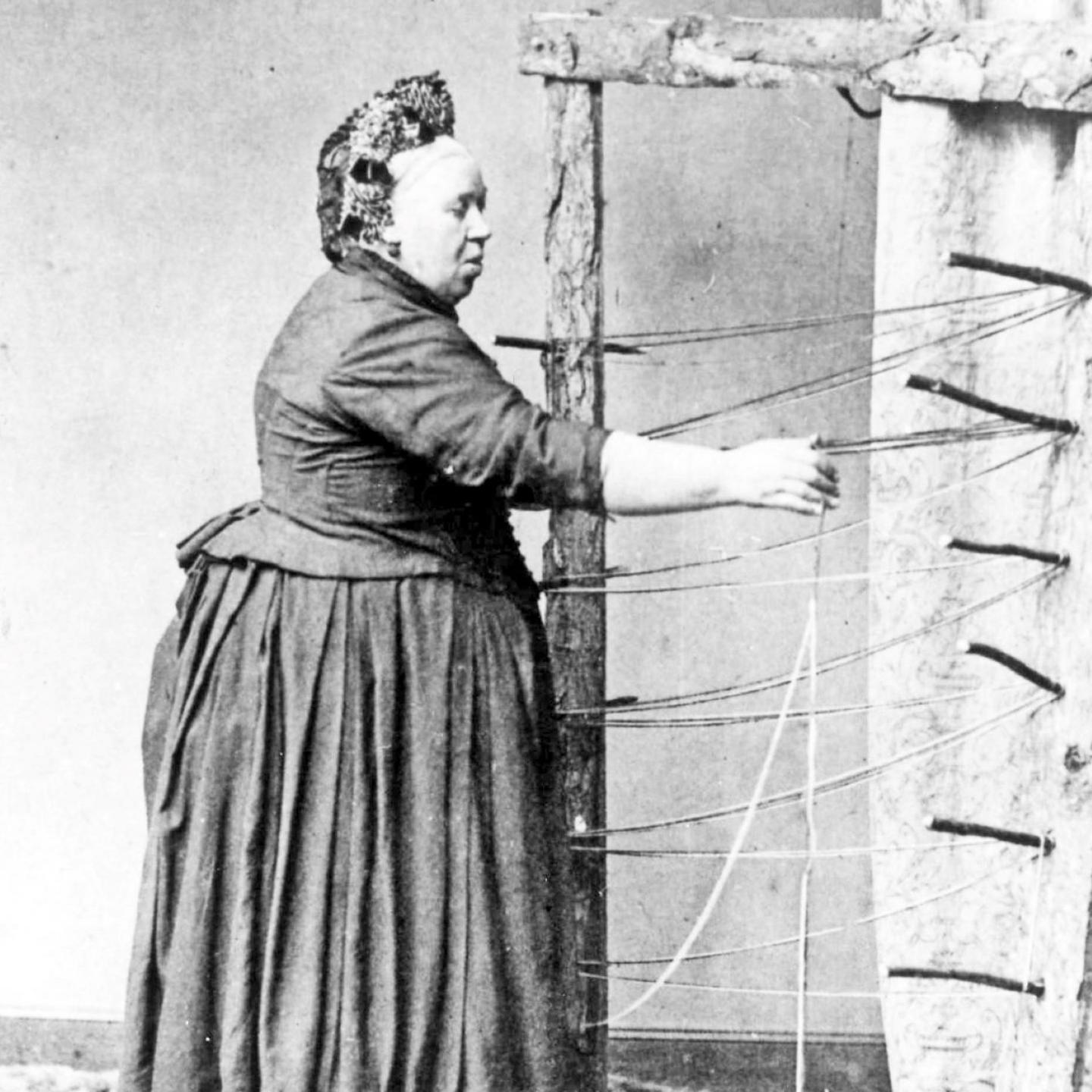 A woman stands weaving threads. She wears Victorian clothing.