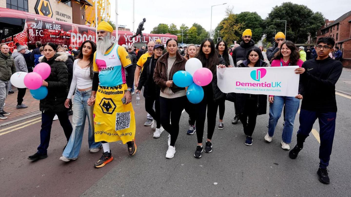 A group of people walking outside a football stadium. Some of the group are holding pink, white and blue balloons.