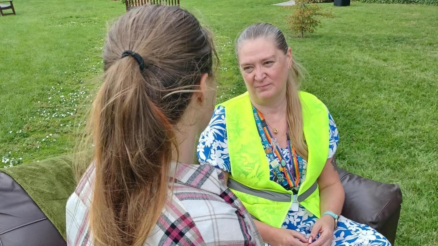 Lisa Pearson with long blonde hair wearing a blue and white dress and a yellow hi-vis vest. She is sitting on a dark leather sofa facing the camera. Another woman with long brown hair is facing her wearing a red and white shirt. The sofa is on grass.