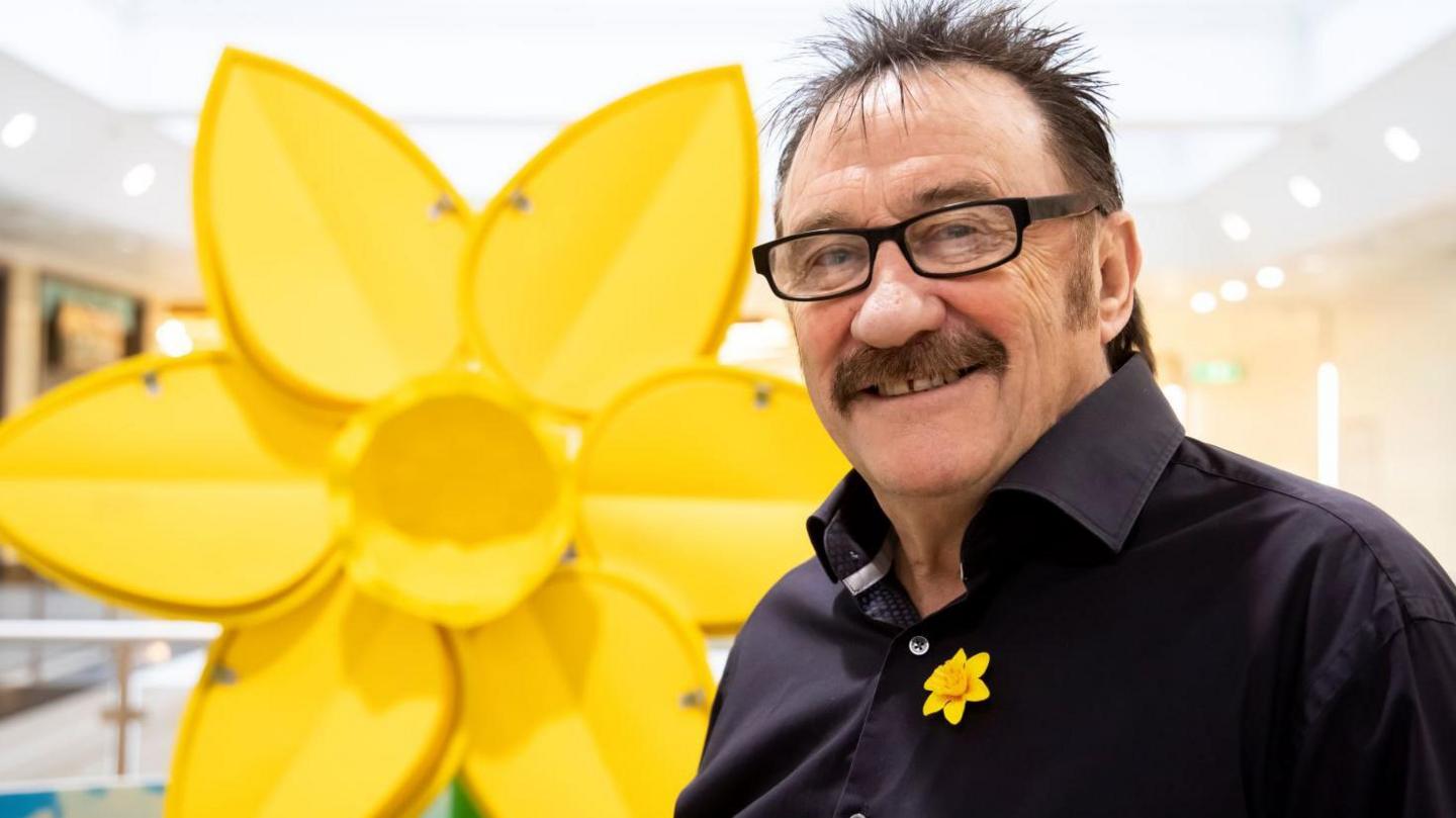 Paul Chuckle smiling looking straight on. He is wearing a black shirt with a yellow daffodil flower badge. He is stood next to a large daffodil-shaped statue. 