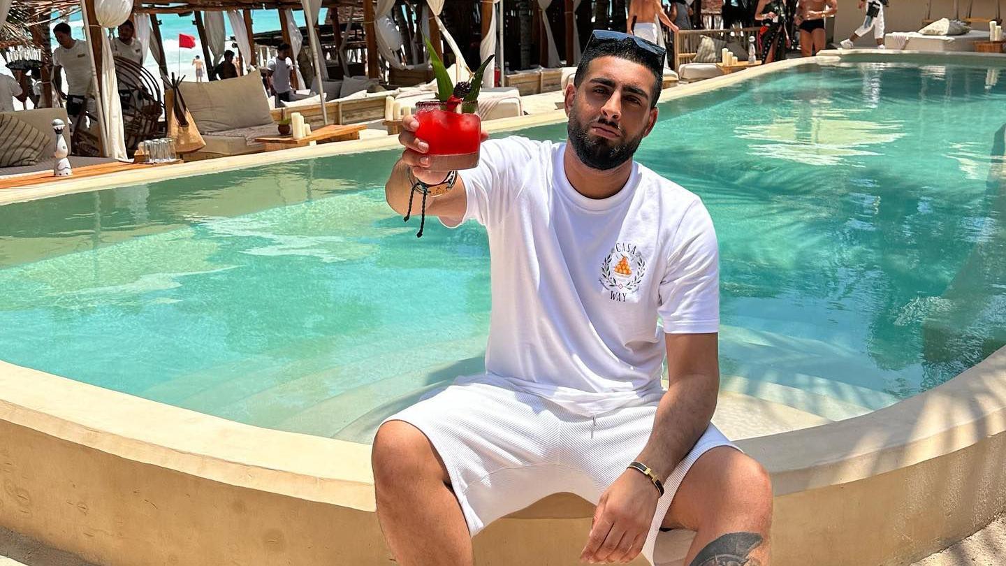 Dyal sits on the edge of a sandstone-coloured pool with turquoise water behind him and sunloungers and tables behind that. He's wearing white T-shirt and shots, sunglasses pushed back on his head and holding up a red-coloured cocktail.