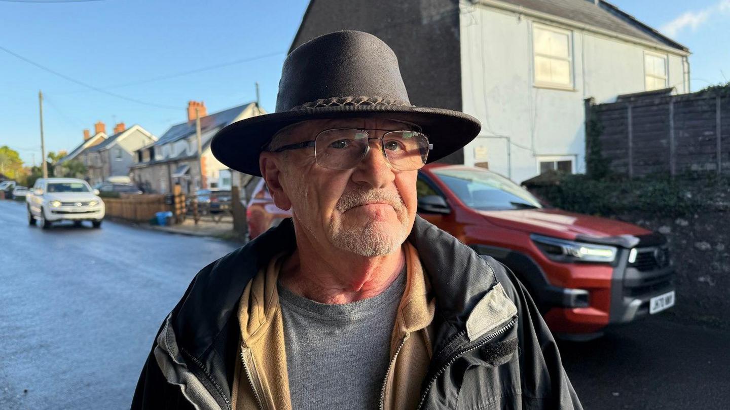 Man wearing hat and glasses looks at camera on side of road with red and white cars behind him
