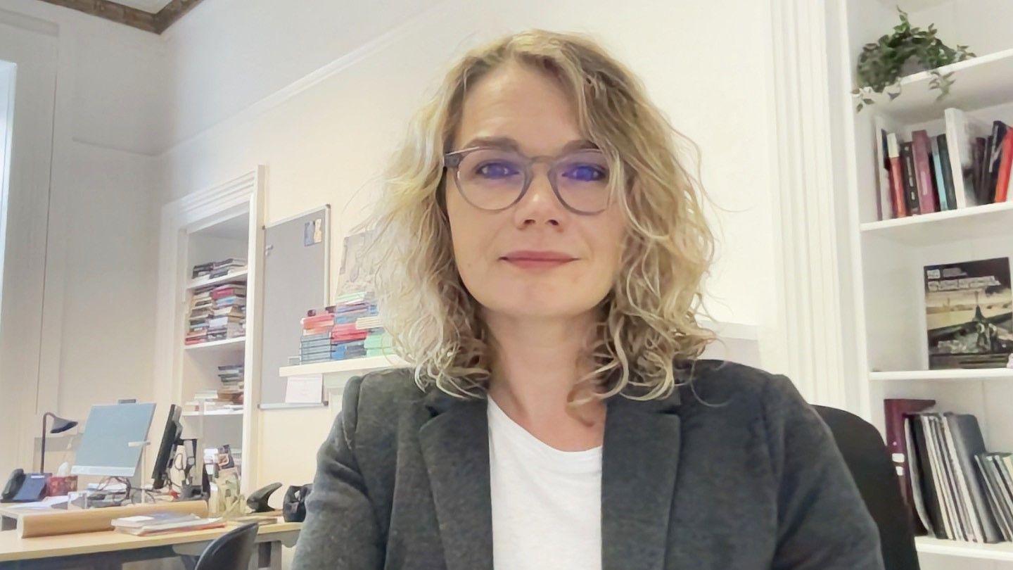 Dr Kelly Johnson, a woman with shoulder-length blonde curly hair and big stylish glasses, looks at the camera while sitting in her office. She wears a white t-shirt and checked wool blazer.