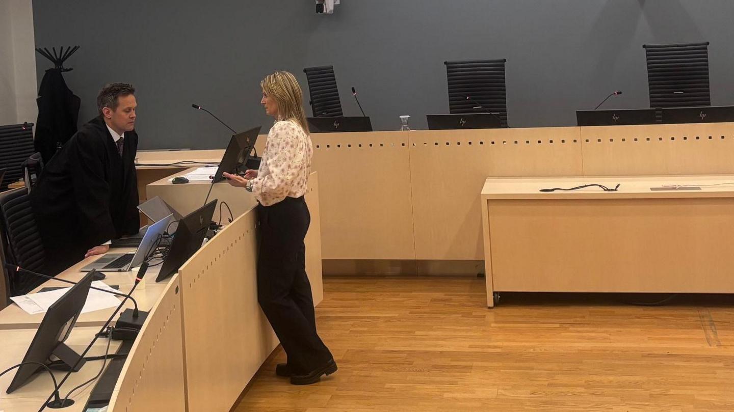 A man to the left talks to a woman in a courtroom - he wears a suit, she wears a light coloured blouse