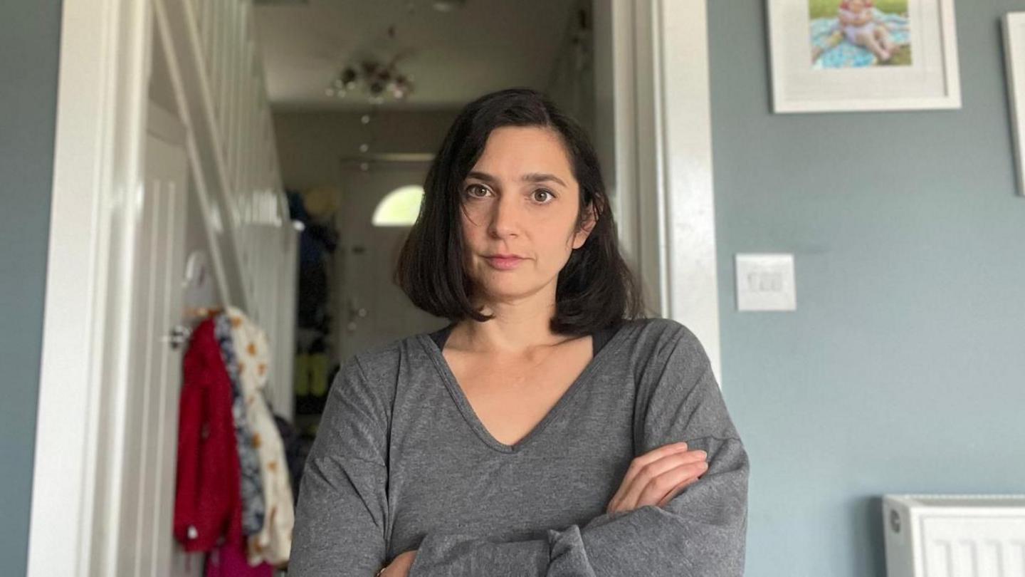 A woman with black hair and a grey jumper with her arms crossed looking seriously at the camera.