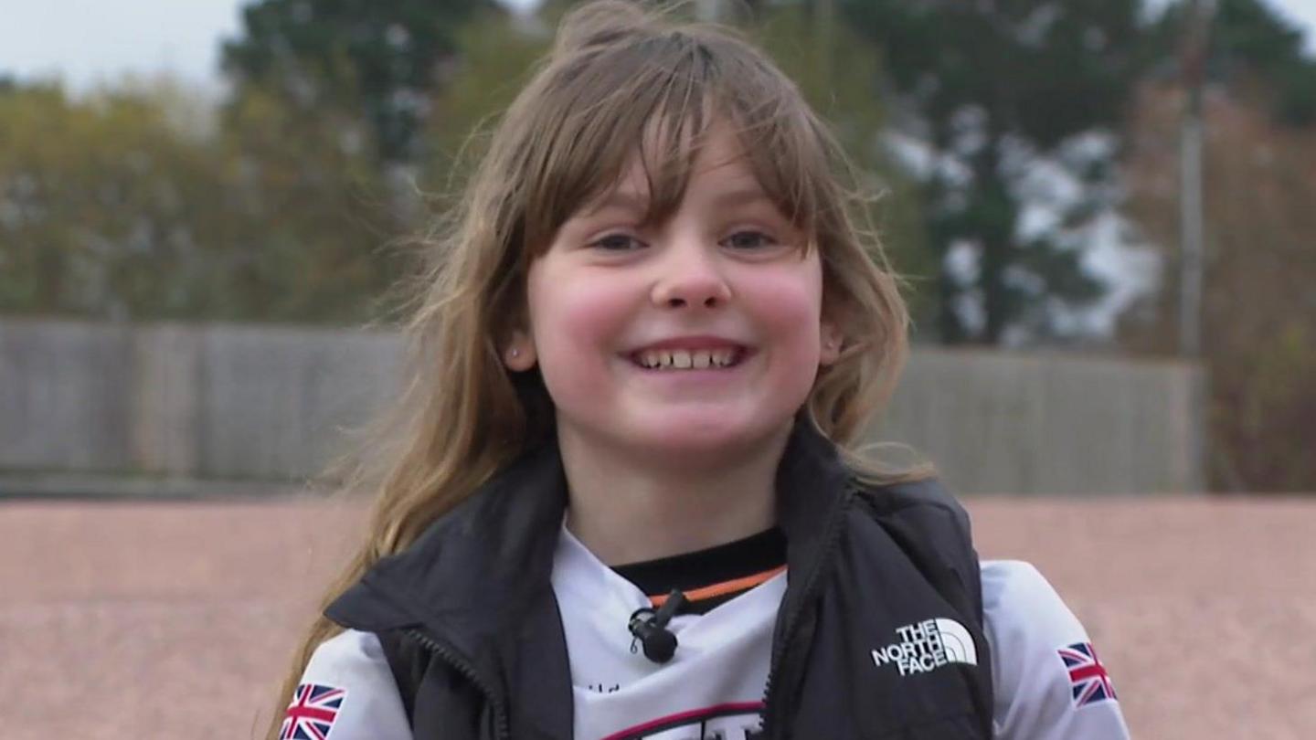 Close up image of Ella smiling at the camera. Ella has long brown hair with a fringe covering her forehead. She is wearing a white Exeter Eagles top with two Union Jacks on each arm. She has a black The North Face gilet on. She is sat on the BMX track. Green trees are in the background.