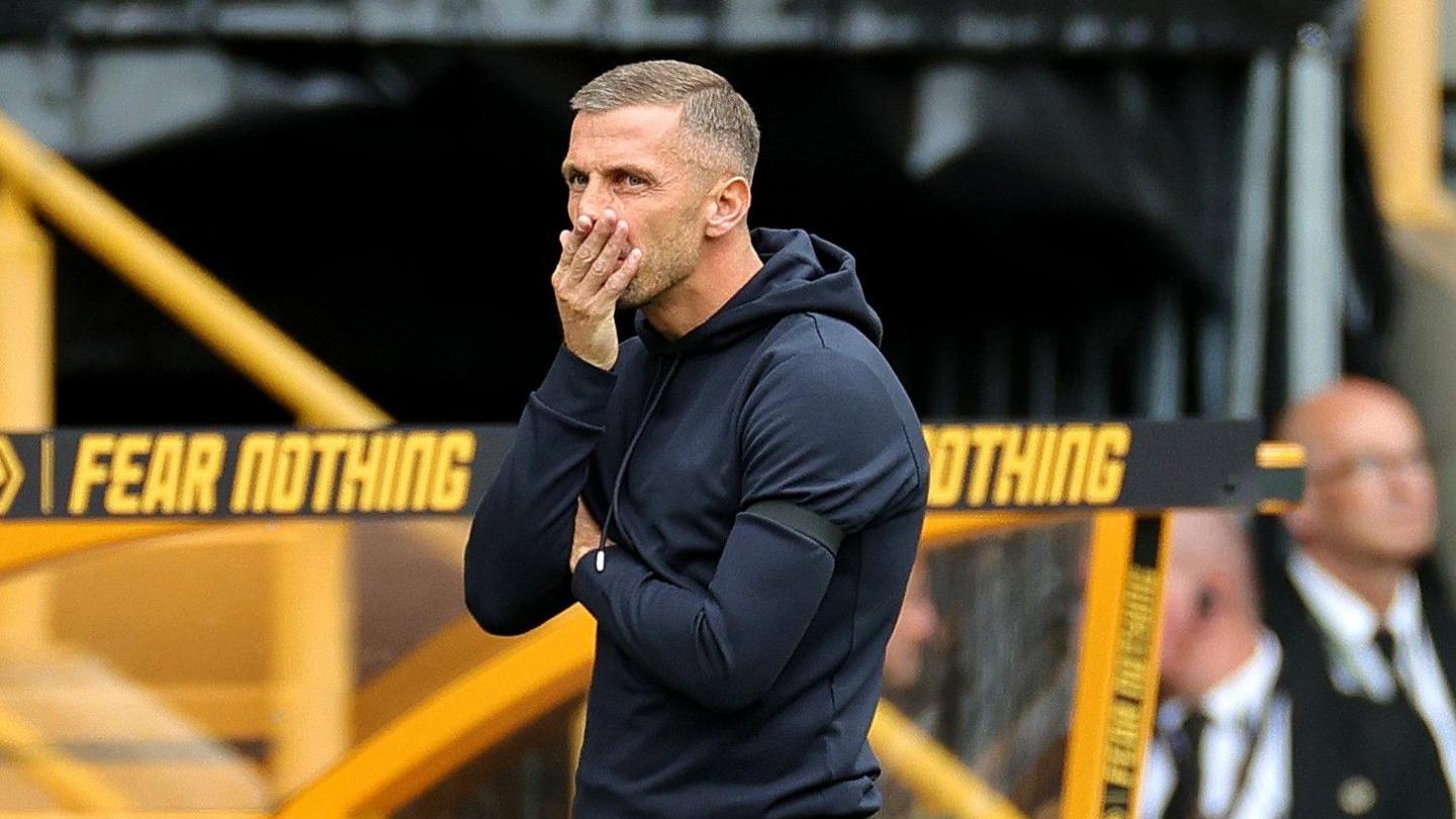 Gary O'Neil, in a black Wolves tracksuit, puts his right hand to his mouth as stands in front of a yellow dugout with the words 'fear nothing' written on the top while watching Wolves' defeat to Chelsea.