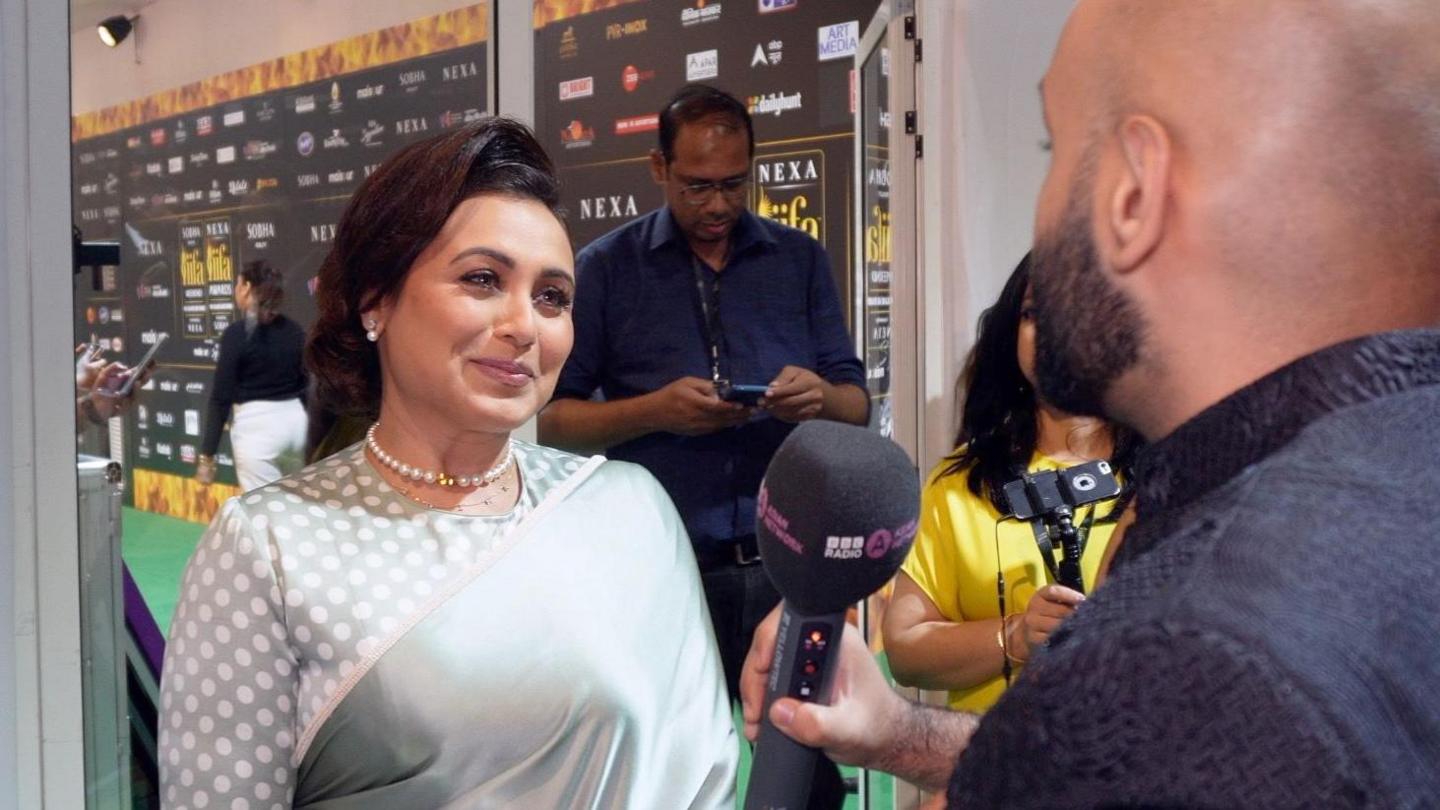 Haroon Rashid, holding a dark microphone, asking Rani Mukerji a question on the IIFA Awards green carpet, with Rani wearing a traditional shiny grey sari and white pearl necklace.