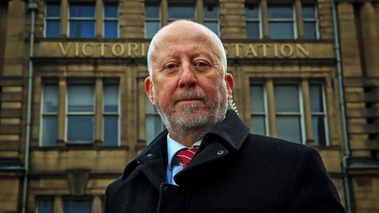 Middlesbrough and Thornaby MP Andy McDonald, wearing a black coat and red striped tie and standing outside Victoria Station, which is a large Victorian stone building with Victoria Station in gold metal letters.