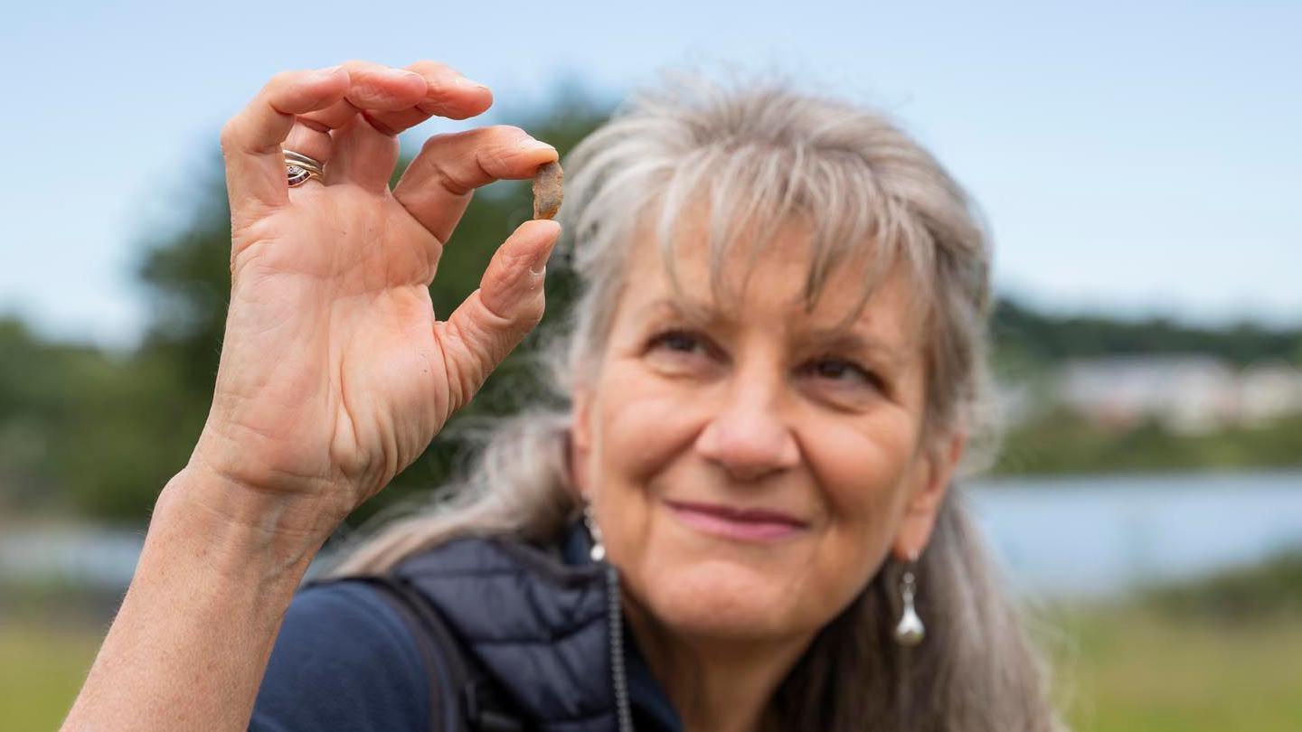 A woman is pictured on site at Sutton Hoo. She is holding a fragment of a historical find in her hand in front of her face. She is looking at the item and smiling. She has long grey hair with a fringe. 