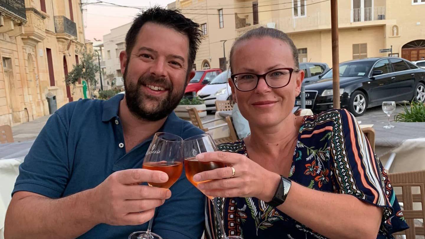 A bearded man in a blue shirt and a blonde woman with spectacles in a black patterned dress toast glasses of rose in an outside restaurant somewhere abroad. There are cars parked behind them in the square.