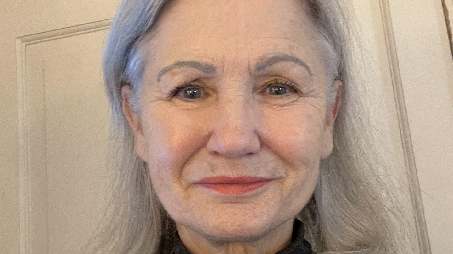 Julie Colville is an older woman with long straight grey hair and blue eyes. He stands smiling in front of a cream-coloured door. 