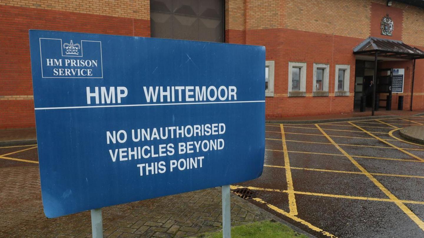 A sign at the entrance to HMP Whitemoor, which is a prison that has large brick walls. The sign is blue with white text, advertising the fact it is HMP Whitemoor and that no unauthorised vehicles should go beyond the sign.