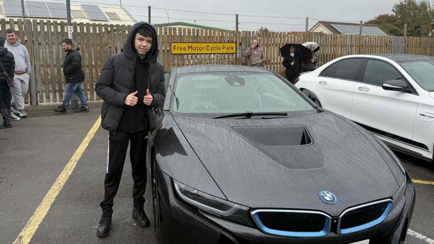 A young teenage boy wearing all black - including a coat with the hood up and jogging bottoms - gives two thumbs up as he stands beside a black BMW in a rainy car park. There is a white car parked beside the BMW and other people can be seen in the background.