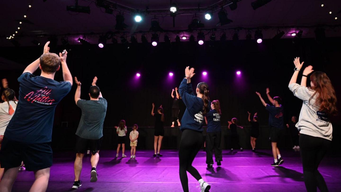 A group of dancers, pictured from the back of the room, shown stepping to the side and clapping above their heads. They are dancing on a dark stage, lit by white and purple lights. The floor is bright purple, lit by the lights. 