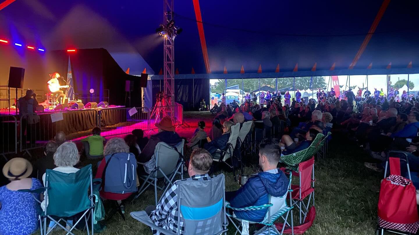 People sat on camping chairs in a large festival marquee watching a man who is sat down playing a guitar on the stage in front of them. The marquee is lit with pink and purple lighting.